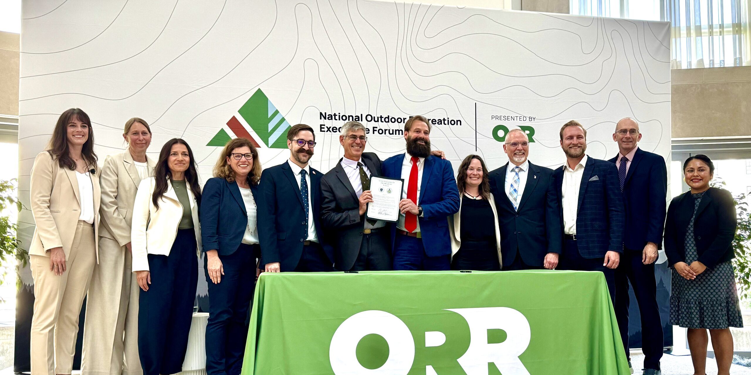 13 people stand smiling behind a table holding up a document