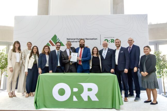 13 people stand smiling behind a table holding up a document