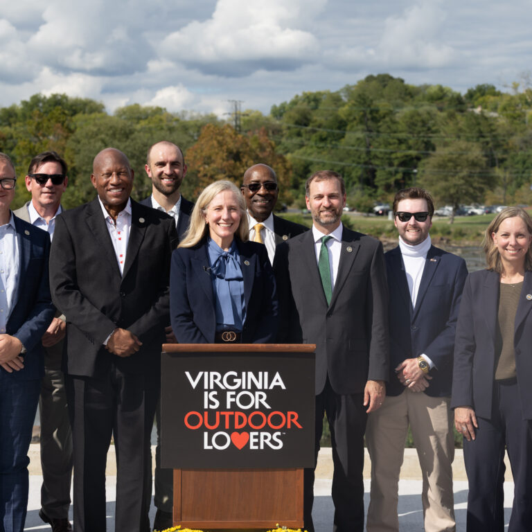 C27A0949 Governor Spanberger and eleven professionals outside, around a podium that reads "Virginia is for Outdoor Lovers."